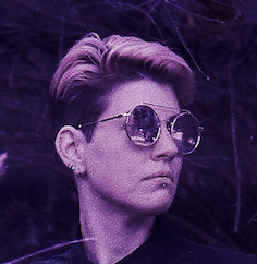 Headshot of author, rendered in shades of purple. They are white, with a two-tone undercut, and are looking to the right with a neutral expression through large circular sunglasses. They have a labret piercing, and several lobe piercings are visible on their right ear.